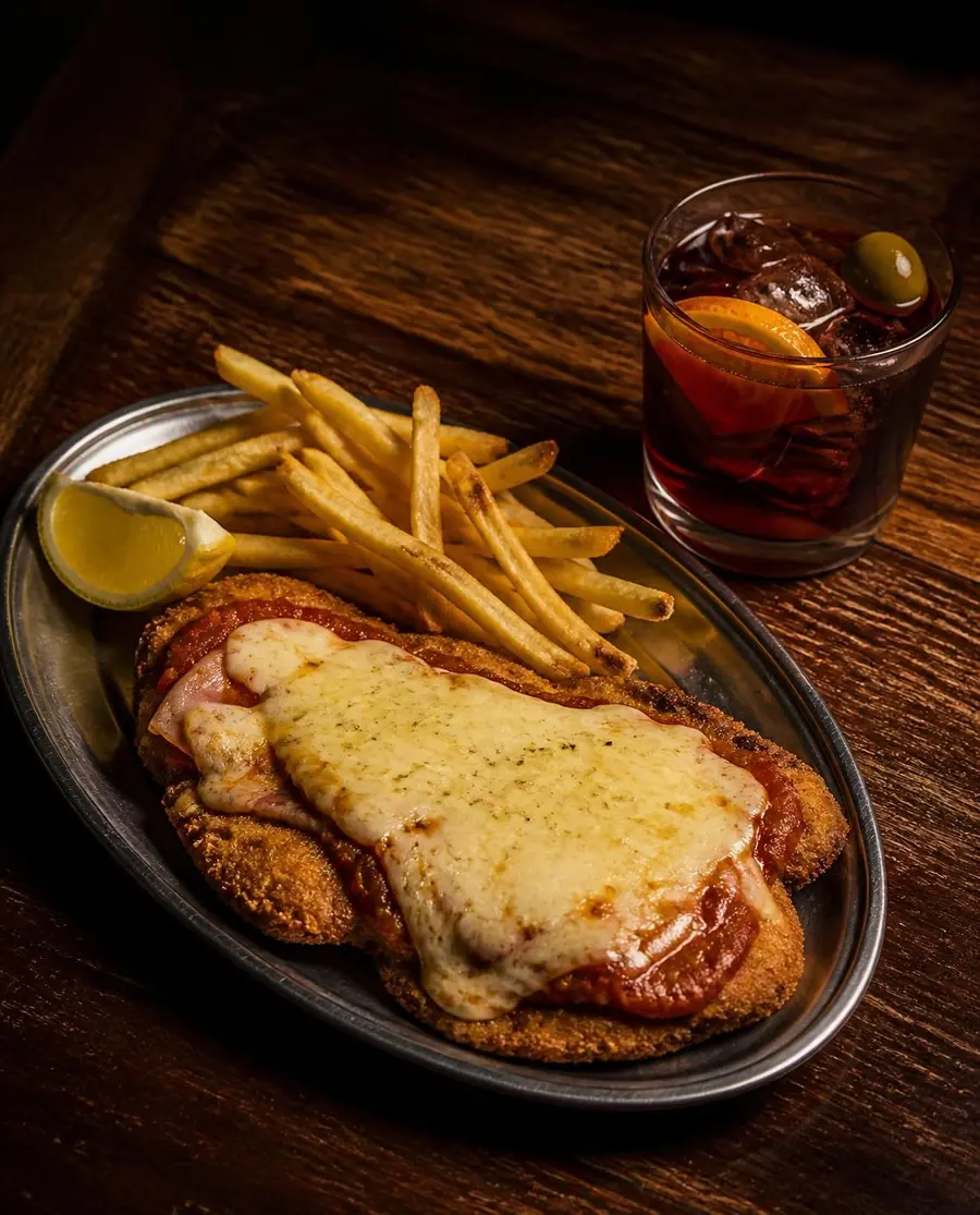Milanesa napolitana con jamón, muzzarella gratinada y salsa, papas fritas y limón, junto a un vermú con naranja y aceituna sobre una mesa de madera